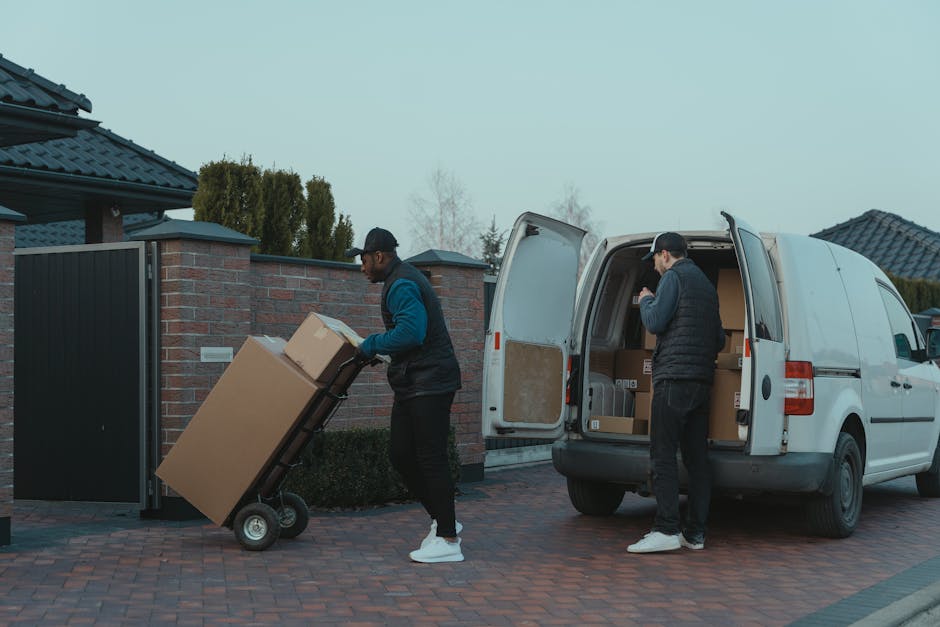 Two movers from Man With a Van Hornchurch are engaged in a home relocation process on a paved driveway in Hornchurch, with a row of residential houses and a brick wall visible in the background. One p