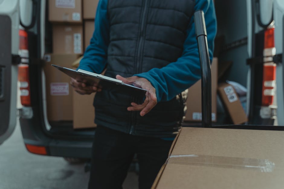 Two movers from Man With a Van Hornchurch are engaged in a home relocation process on a paved driveway in Hornchurch, with a row of residential houses and a brick wall visible in the background. One p