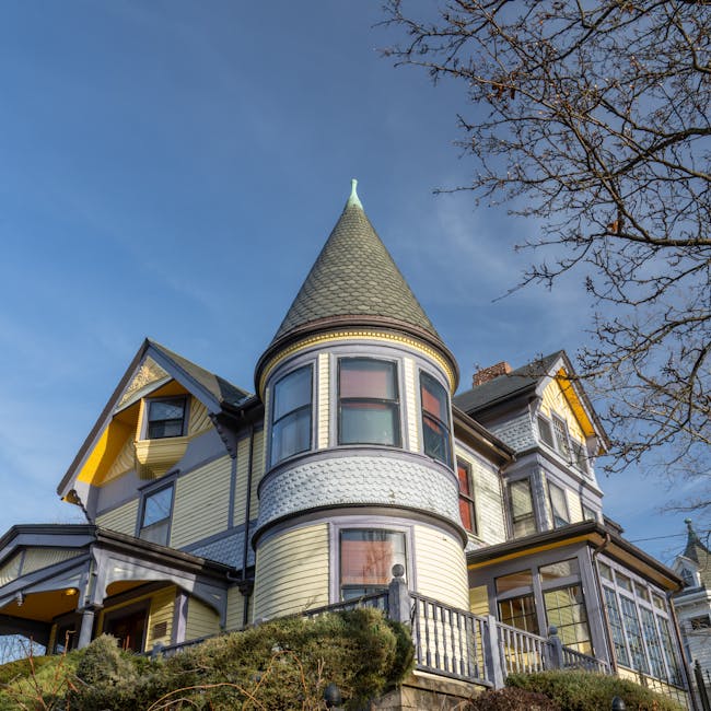 A large Victorian-style house with a distinctive rounded turret featuring multiple windows, situated on a raised garden plot with a wooden fence and steps leading up to the entrance. The house's exterior is clad in light yellow and grey siding, with decorative trim and gabled roof sections, including a prominent conical tower roof. The surrounding environment includes a clear blue sky, a leafless tree on the right side, and well-maintained shrubbery at the front. This property is located in Hornchurch, and the image relates to house removals and relocations, highlighting the importance of careful packing and transport of furniture and other household items during moving services provided by Man With a Van Hornchurch.