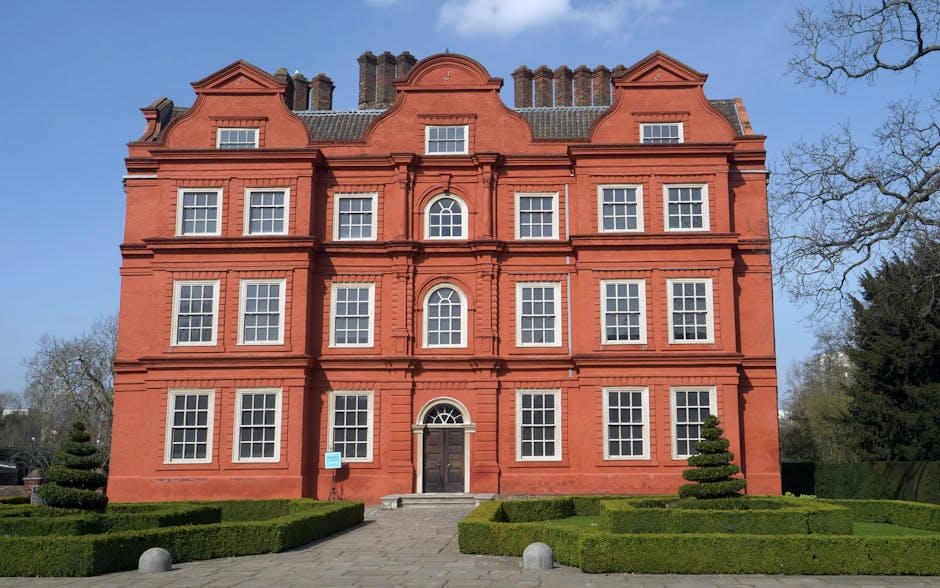 The image shows a historic, multi-storey red-brick building with classical architectural details, including decorative window frames and a distinctive curved gable roof. The building is situated in a well-maintained garden with neatly trimmed hedges and small, rounded topiary trees positioned symmetrically on either side of the central entrance. The entrance features a dark wooden door with a semi-circular fanlight window above, accessible via a small set of steps. The windows are uniformly arranged across the facade, with white frames contrasting against the red brickwork. The surroundings include clear blue sky and leafless trees, indicating a winter or early spring season. This setting exemplifies the type of period property often involved in home relocation or furniture transport projects. As part of professional house removals, tools such as trolleys or moving blankets may be used within or nearby the building for packing and loading purposes. Man With a Van Hornchurch occasionally performs such house removals, ensuring careful handling of furniture and possessions during the loading process.