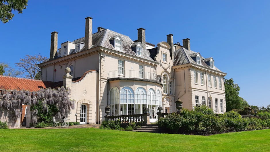 A large Victorian-style house with a distinctive rounded turret featuring multiple windows, situated on a raised garden plot with a wooden fence and steps leading up to the entrance. The house's exterior is clad in light yellow and grey siding, with decorative trim and gabled roof sections, including a prominent conical tower roof. The surrounding environment includes a clear blue sky, a leafless tree on the right side, and well-maintained shrubbery at the front. This property is located in Hornchurch, and the image relates to house removals and relocations, highlighting the importance of careful packing and transport of furniture and other household items during moving services provided by Man With a Van Hornchurch.