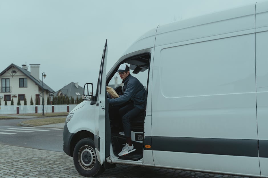 A man wearing glasses, a dark jacket, black trousers, white sneakers, and a black and white cap is sitting inside the cab of a white delivery van with the driver's side door open. He appears to be org