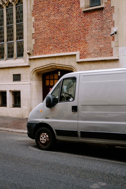 A man wearing glasses, a dark jacket, black trousers, white sneakers, and a black and white cap is sitting inside the cab of a white delivery van with the driver's side door open. He appears to be org