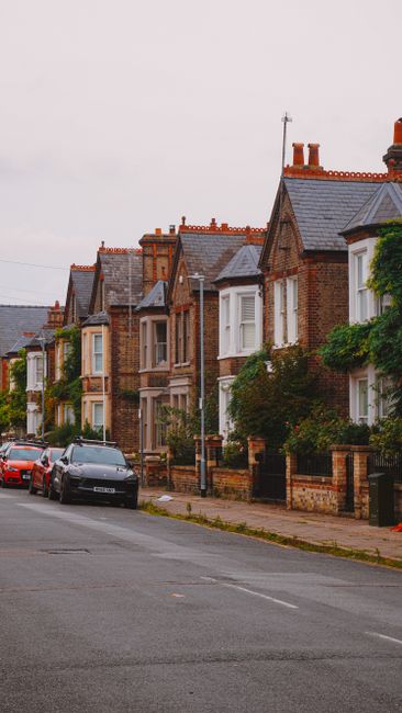 A street view of a residential area with a row of traditional brick houses, each featuring pitched roofs with slate tiles, chimney stacks, and bay windows. Some houses have semi-private front gardens 