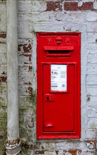 A red British post box mounted on a wooden post and surrounded by green ivy and foliage. The post box features a rectangular slot for mailing letters at the top, with embossed markings including 'POST